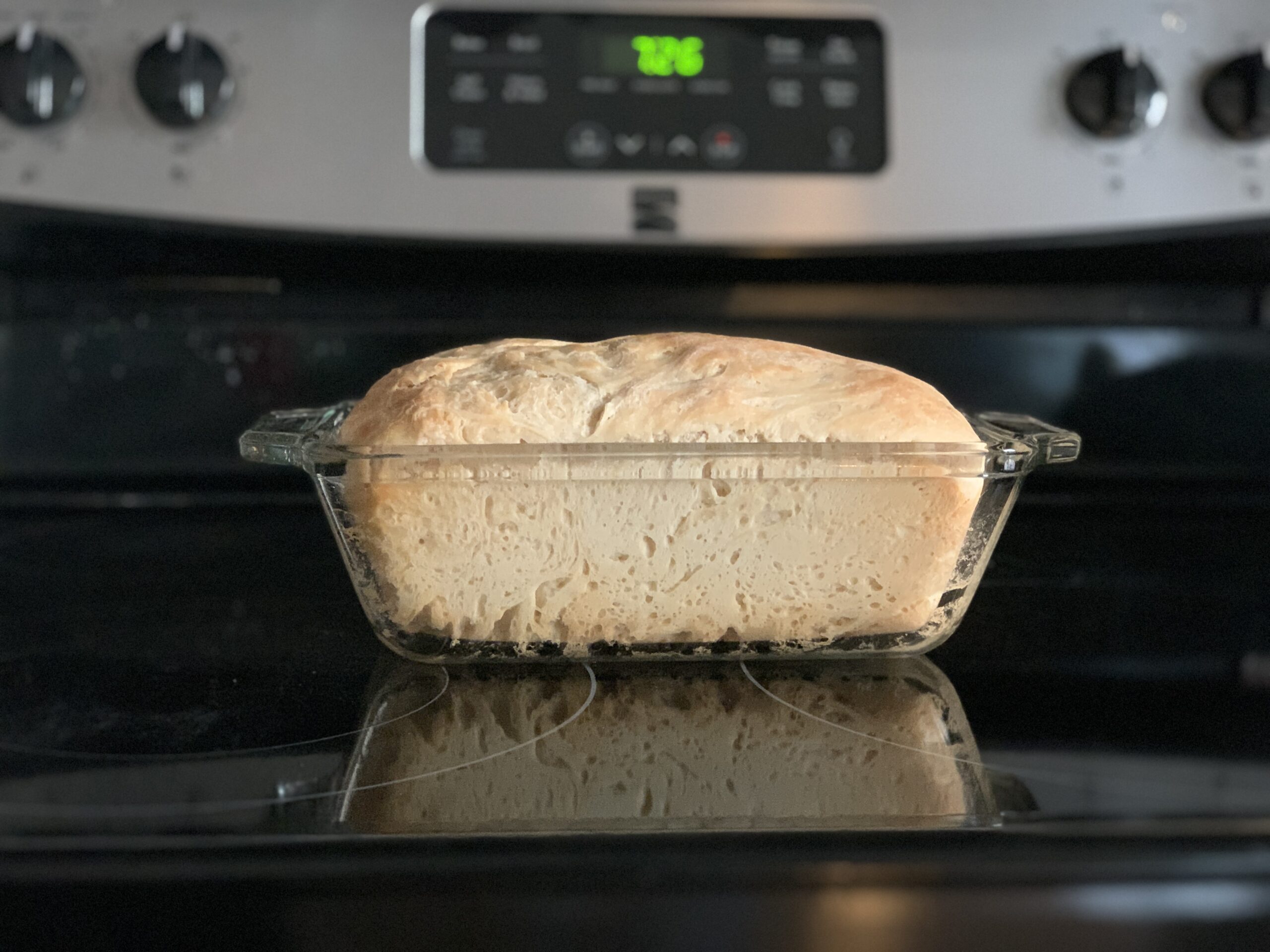 bread in a loaf pan sitting on a black stove top with the time in green on the digital clock in the background