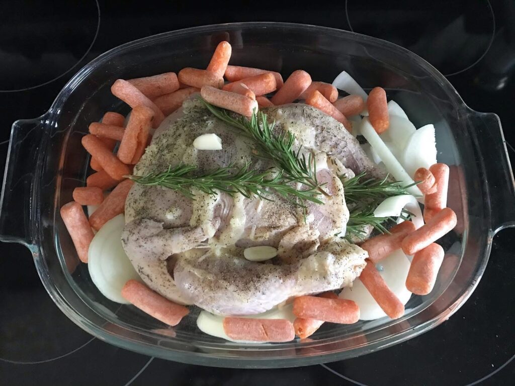 raw whole chicken in a clear roasting pan covered in onions, carrots, and fresh herbs ready to be roasted
