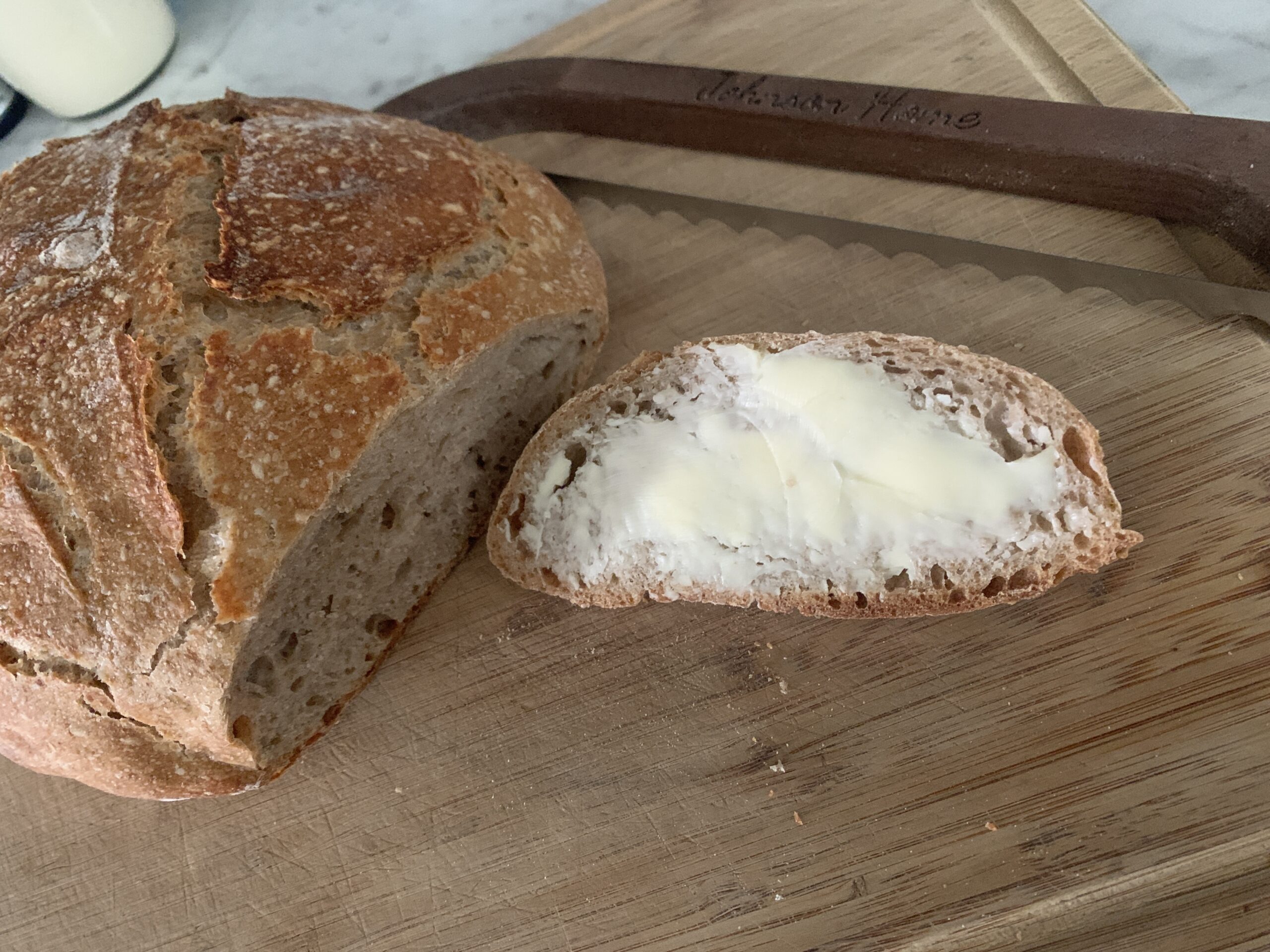 a round artisan sourdough boule of bread cut with one slice buttered on a wooden cutting board with a bread bow in the background