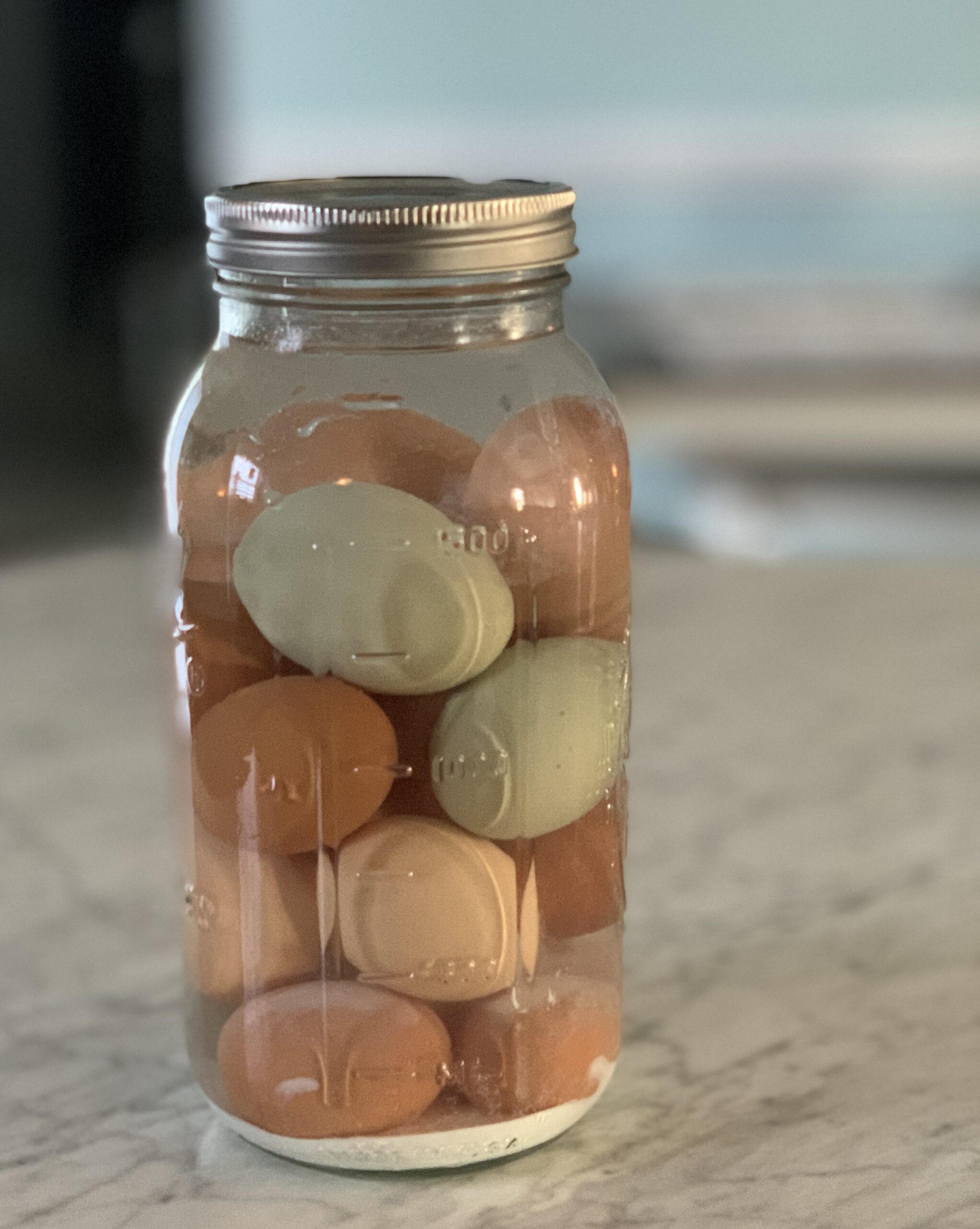 half gallon mason jar full of brown and blue eggs in water with pickling lime settled at the bottom of the jar on a marble counter top