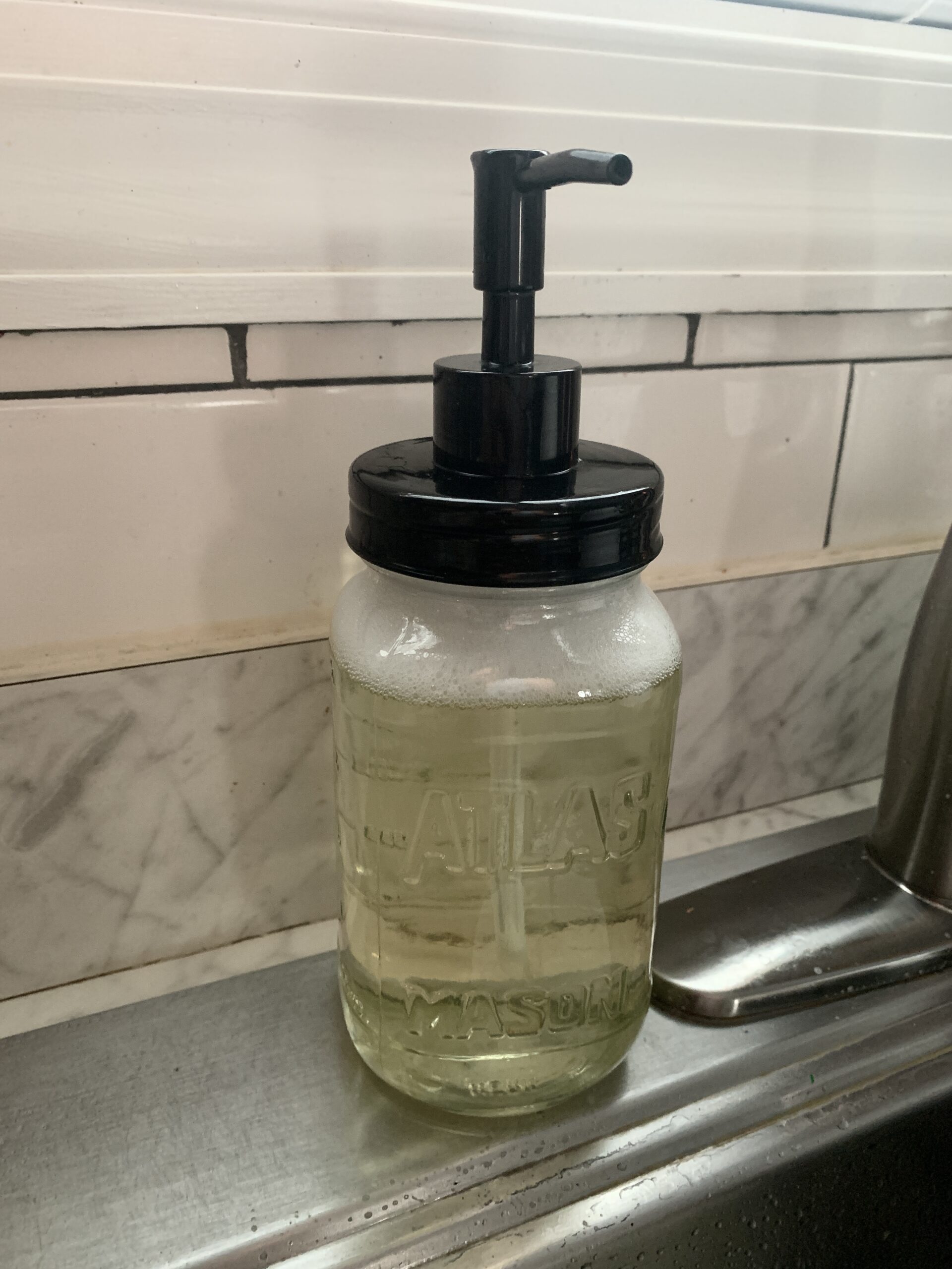 mason jar of DIY dish soap with a black pump top sitting beside a sink faucet