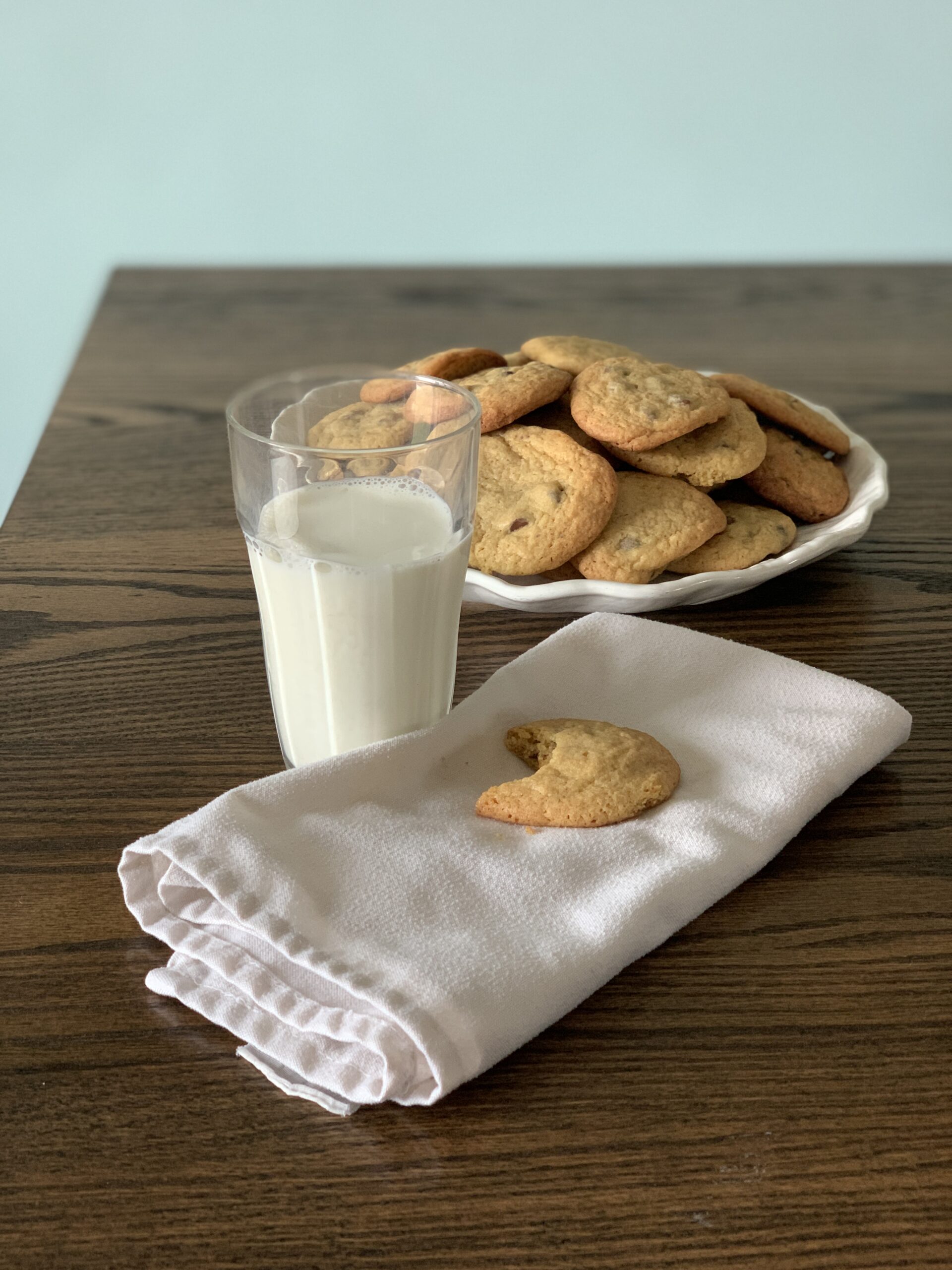 glass of milk and half eaten cookie sits on a white cloth napkin in front of a white round plate of chocolate chip cookies