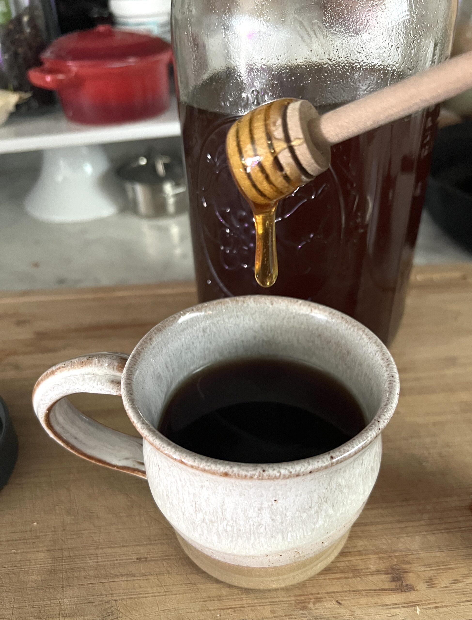 a white pottery mug on a wooden cutting board filled with elderberry tea and a honey spoon dripping honey into the mug