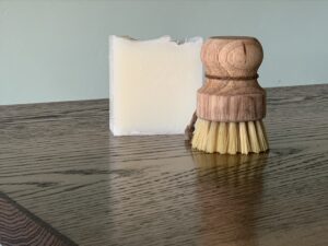 a white square of dish soap bar sitting on a dark wooden table with a dish brush sitting upright in front of the bar of soap
