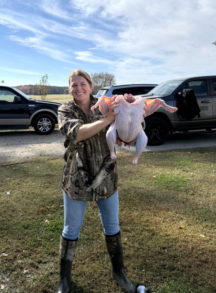 woman in camouflage shirt with jeans tucked into mud boots standing on a sunny day in a yard holding a freshly processed turkey