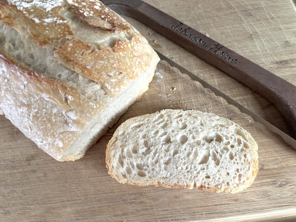 no-fuss sourdough loaf of bread resting on a wooden cutting board with a bread bow knife in the background