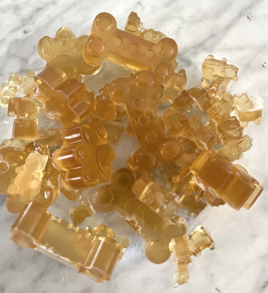 golden colored solid homemade gummy candies on a marble counter top in bear, dog treat bone, and worm shapes