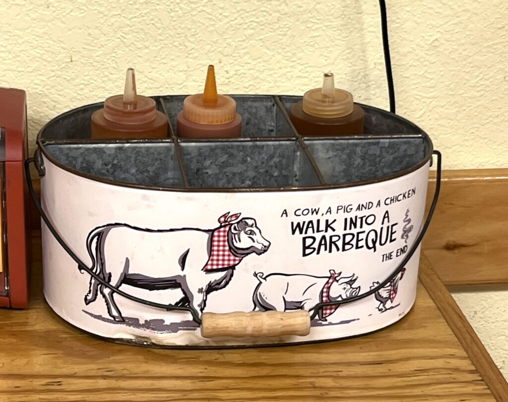 metal container holding 3 bbq sauces that says "a cow, a pig, and a chicken walk into a barbecue, the end."