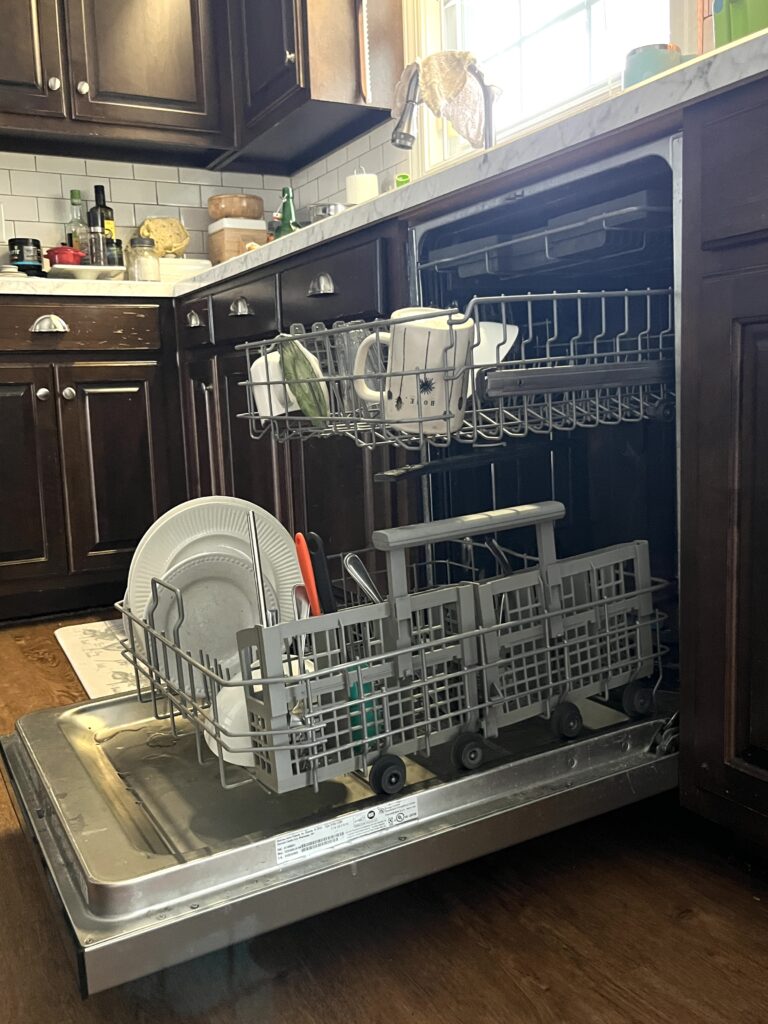 dishwasher opened with racks out and holding several unwashed dishes in a dark brown cabineted kitchen with marble counter tops are a daily cleaning chore