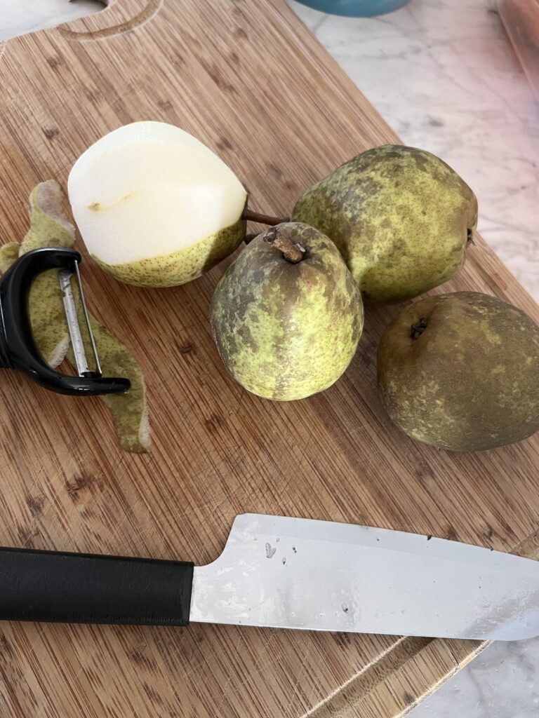 three whole pears and one sliced in half sitting on a wooden cutting board on a marble counter top with a peeler and knife in the foreground