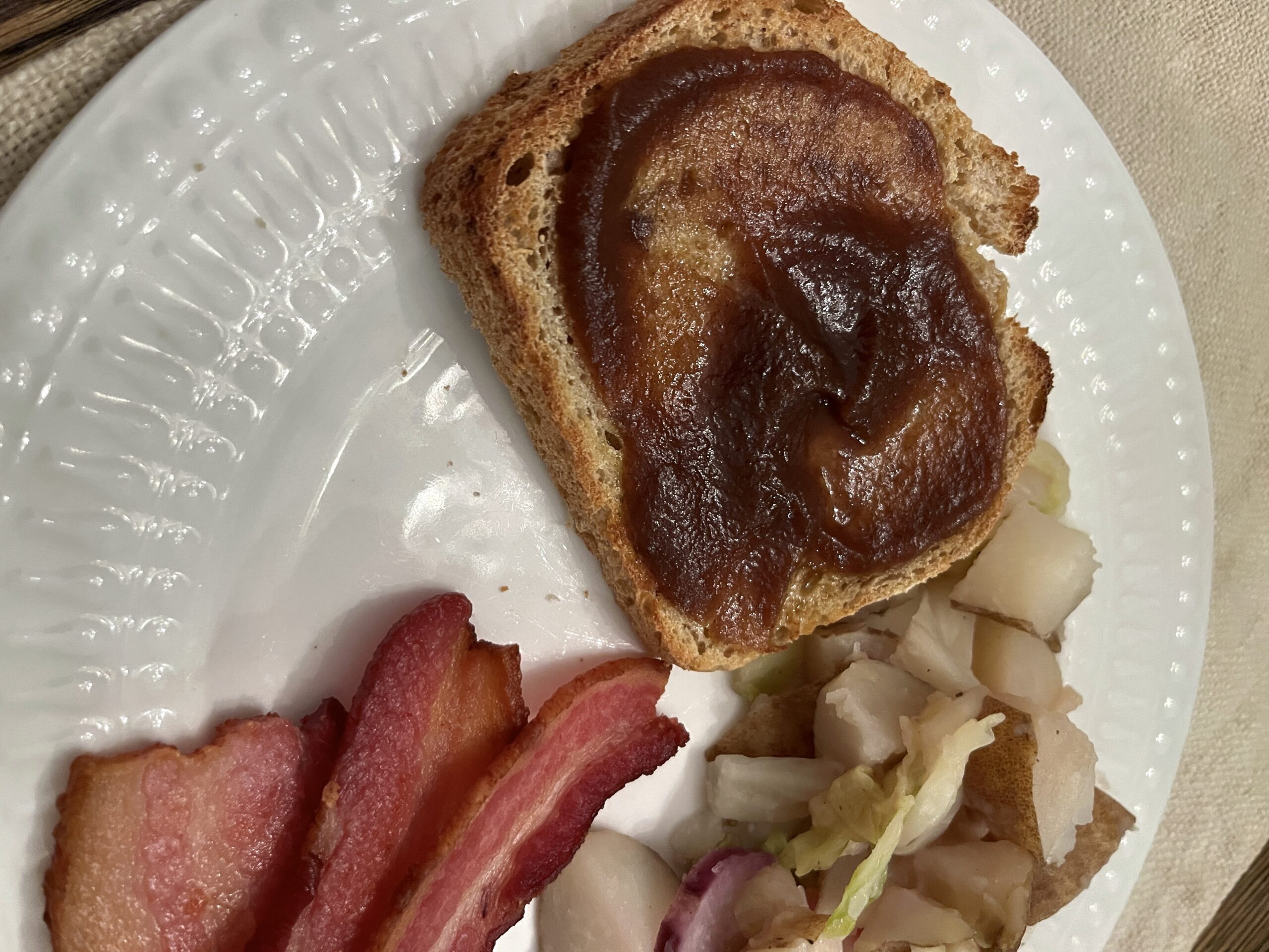 white plate holding thick cut slices of bacon, pan fried potato cubes, and sourdough toast dolloped with rich brown apple butter