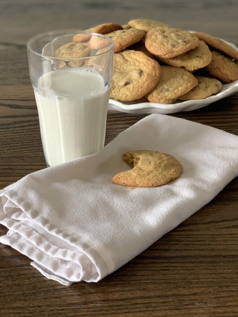glass of milk and half eaten cookie sits on a white cloth napkin in front of a white round plate of chocolate chip cookies