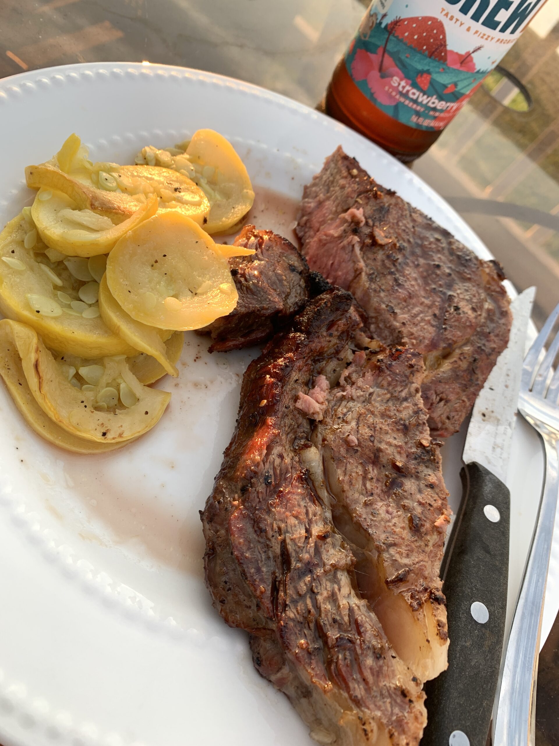 grilled ribeye seasoned to perfection on a white plate accompanied by roasted yellow squash and a kombucha bottle