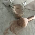 puffy blush brush laying on a white counter spilling diy dry shampoo on to the counter in front of a jar of light brown powder