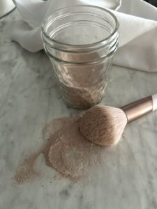 puffy blush brush laying on a white counter spilling diy dry shampoo on to the counter in front of a jar of light brown powder