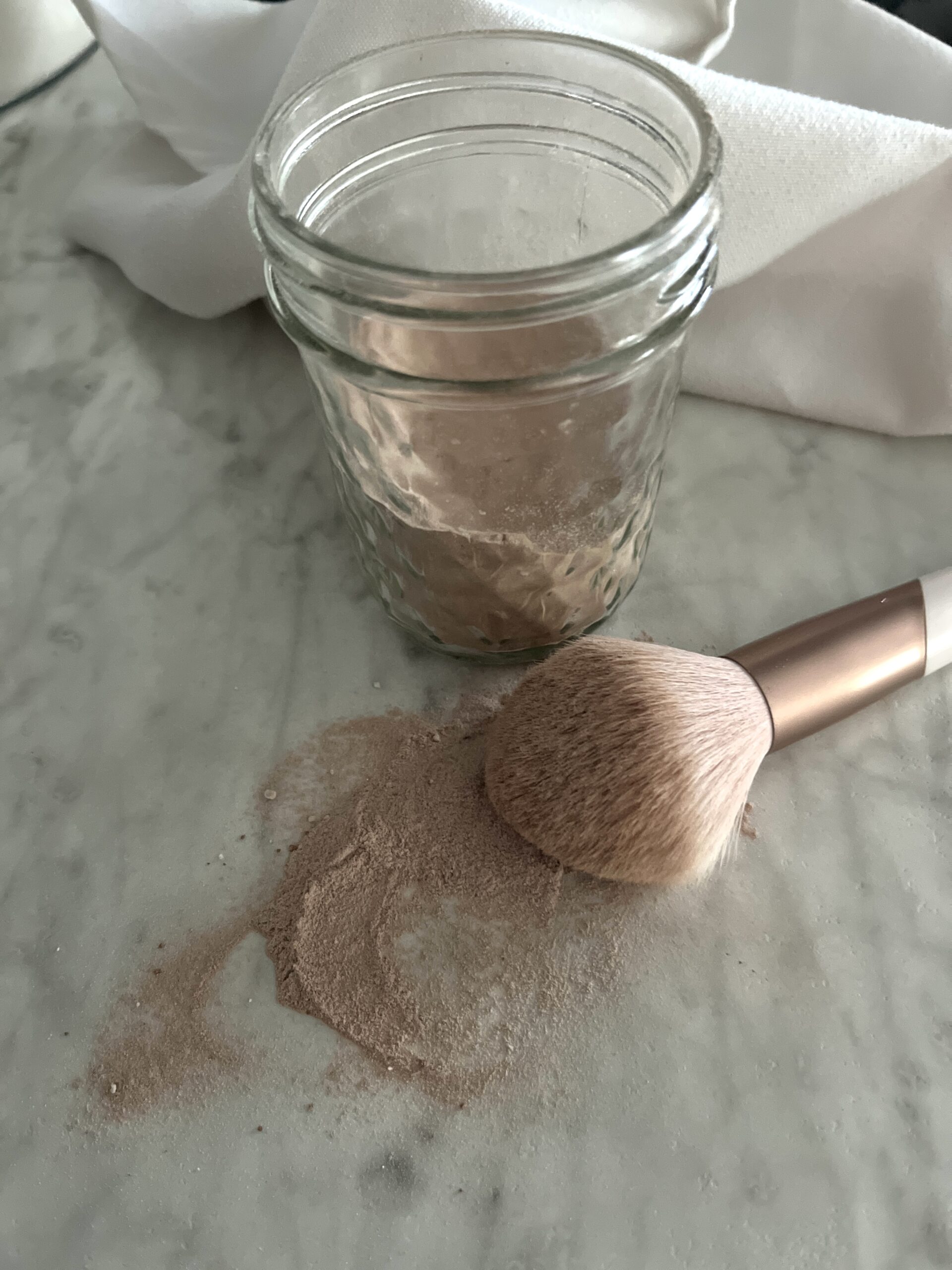 puffy blush brush laying on a white counter spilling diy dry shampoo on to the counter in front of a jar of light brown powder