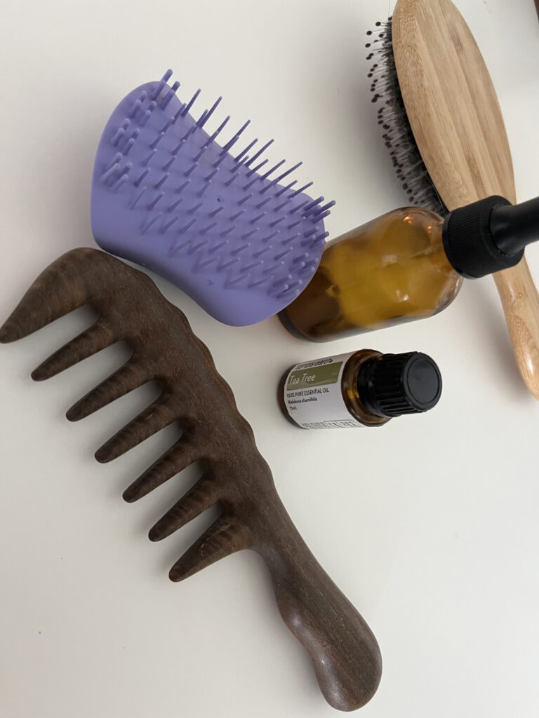 white counter hosting a sandalwood comb, purple scalp brush, amber dropper bottle, Simply Earth tea tree essential oil, and a wooden hair brush