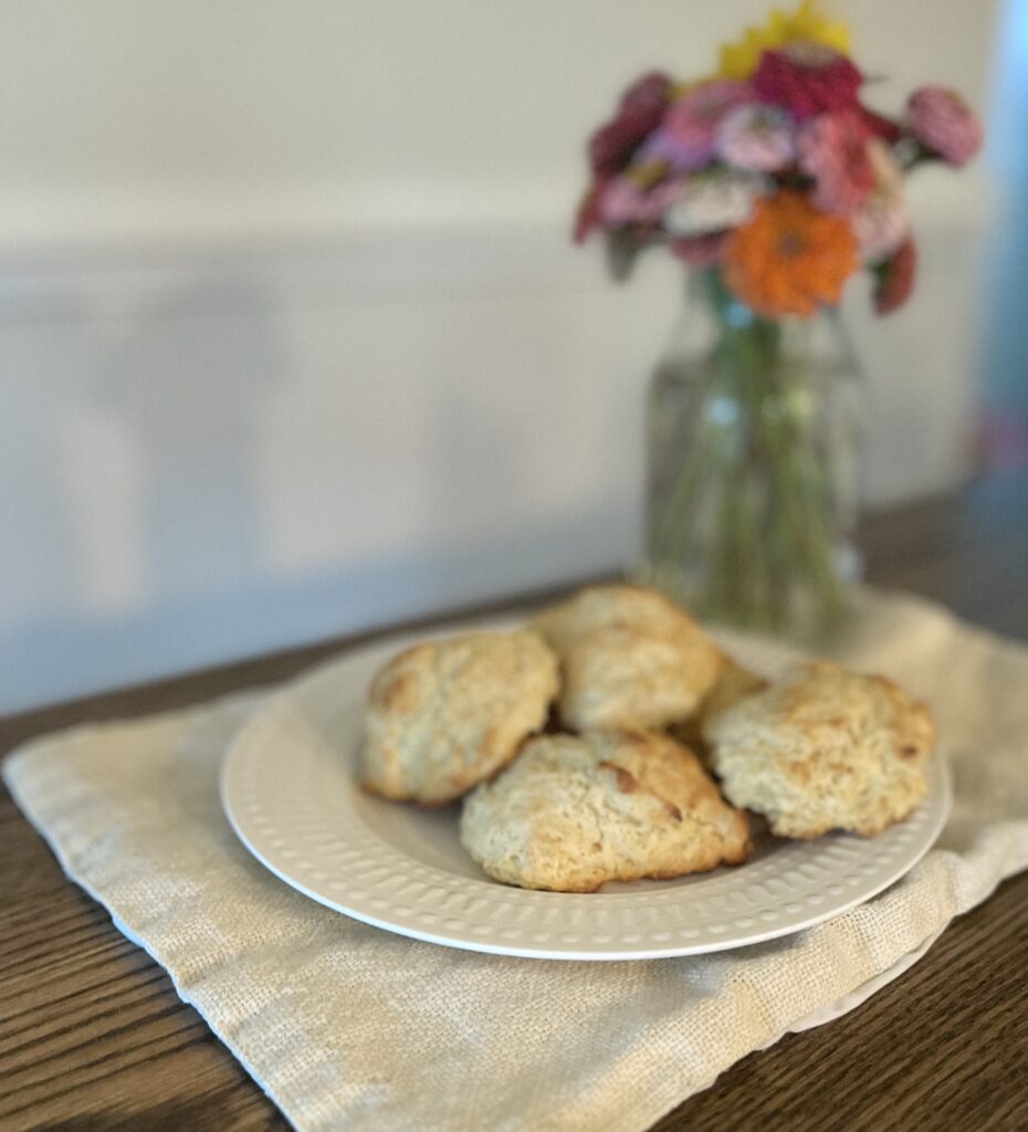 white plate of from scratch drop biscuits sitting on a linen napkin on a wooden table in front of a vase of colorful flowers with a shadow cast on the wall behind
