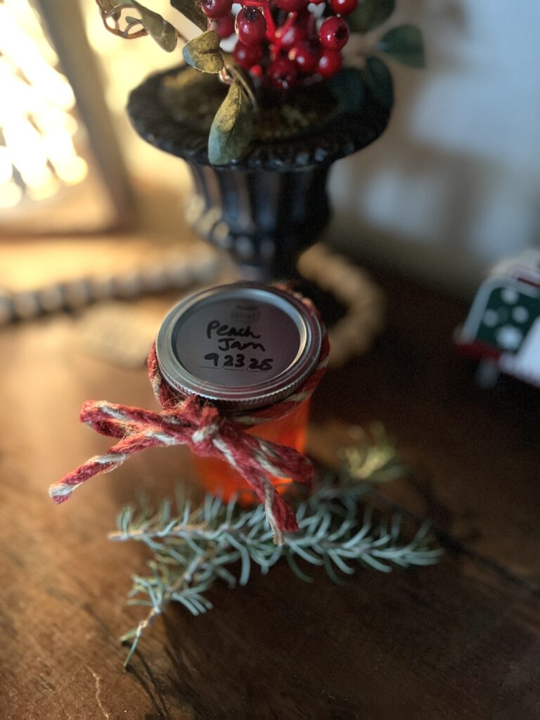 jar of peach jam wrapped in red and brown twine on a wooden table with two sprigs of rosemary lying in front of it. Light being cast from the left of the jar.
