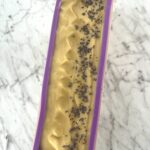 a loaf of goats milk soap with lavender buds on top poured into a purple soap mold sitting on a white marble counter top
