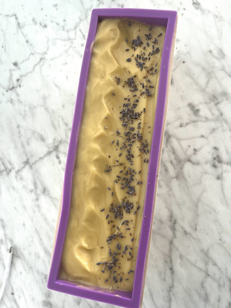 a loaf of goats milk soap with lavender buds on top poured into a purple soap mold sitting on a white marble counter top