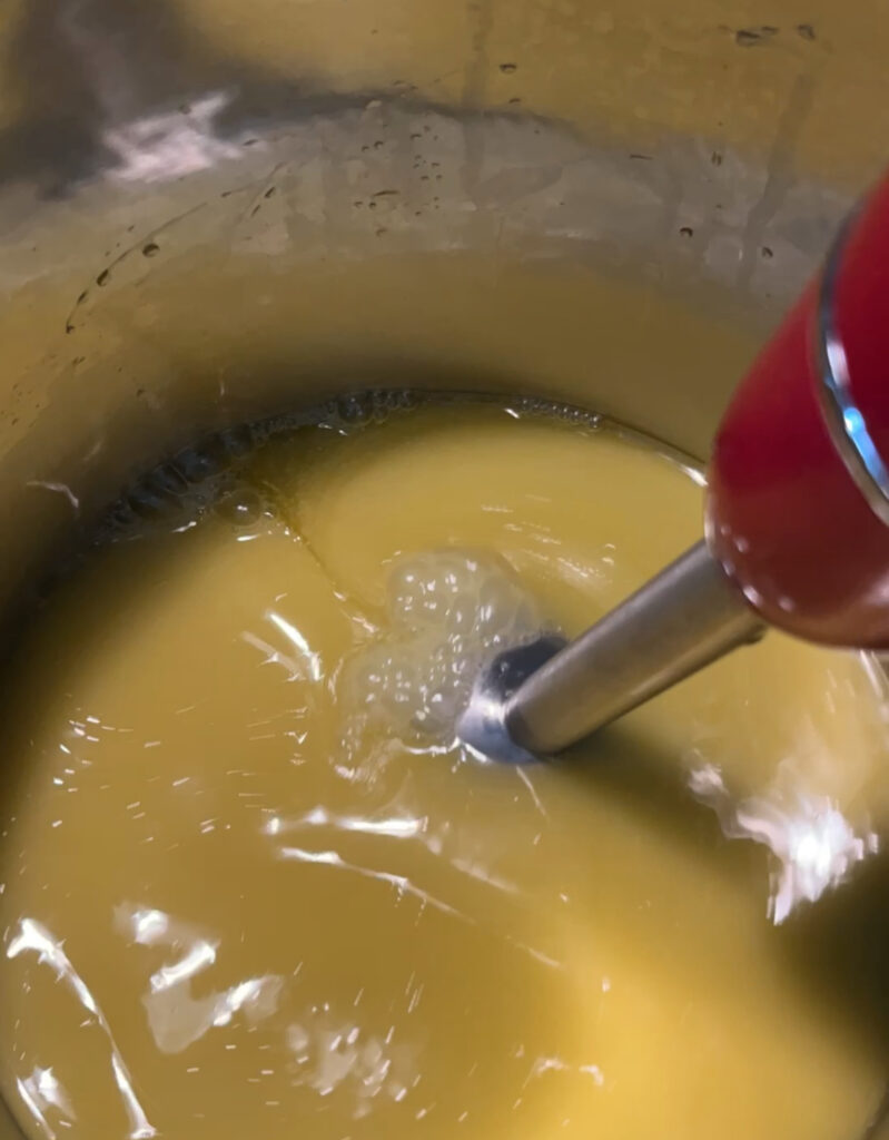 yellow colored soap being mixed in a silver stock pot with a red immersion stick blender