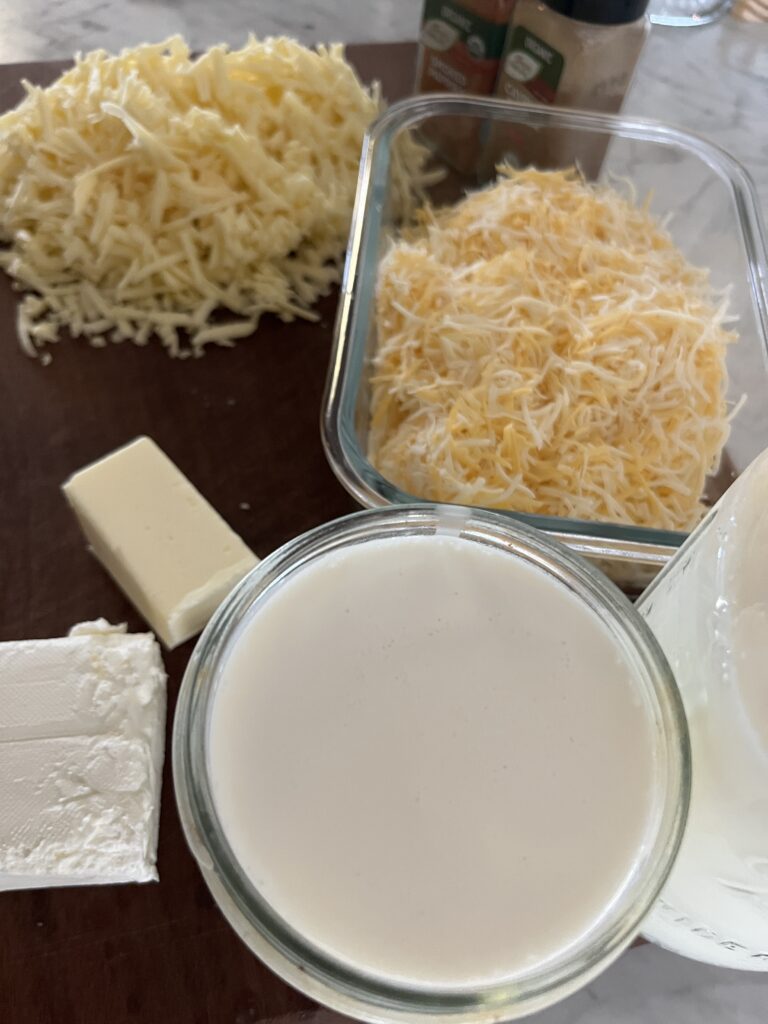 one pile of white shredded cheese in the forefront with a glass dish of yellow and white cheese in the background accompanied by butter, cream cheese, and two jars of milk on a dark wooden cutting board