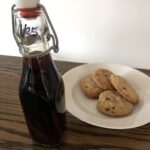 glass swing top bottle of vanilla extract sitting on wooden table in front of a small white plate of chocolate chip cookies
