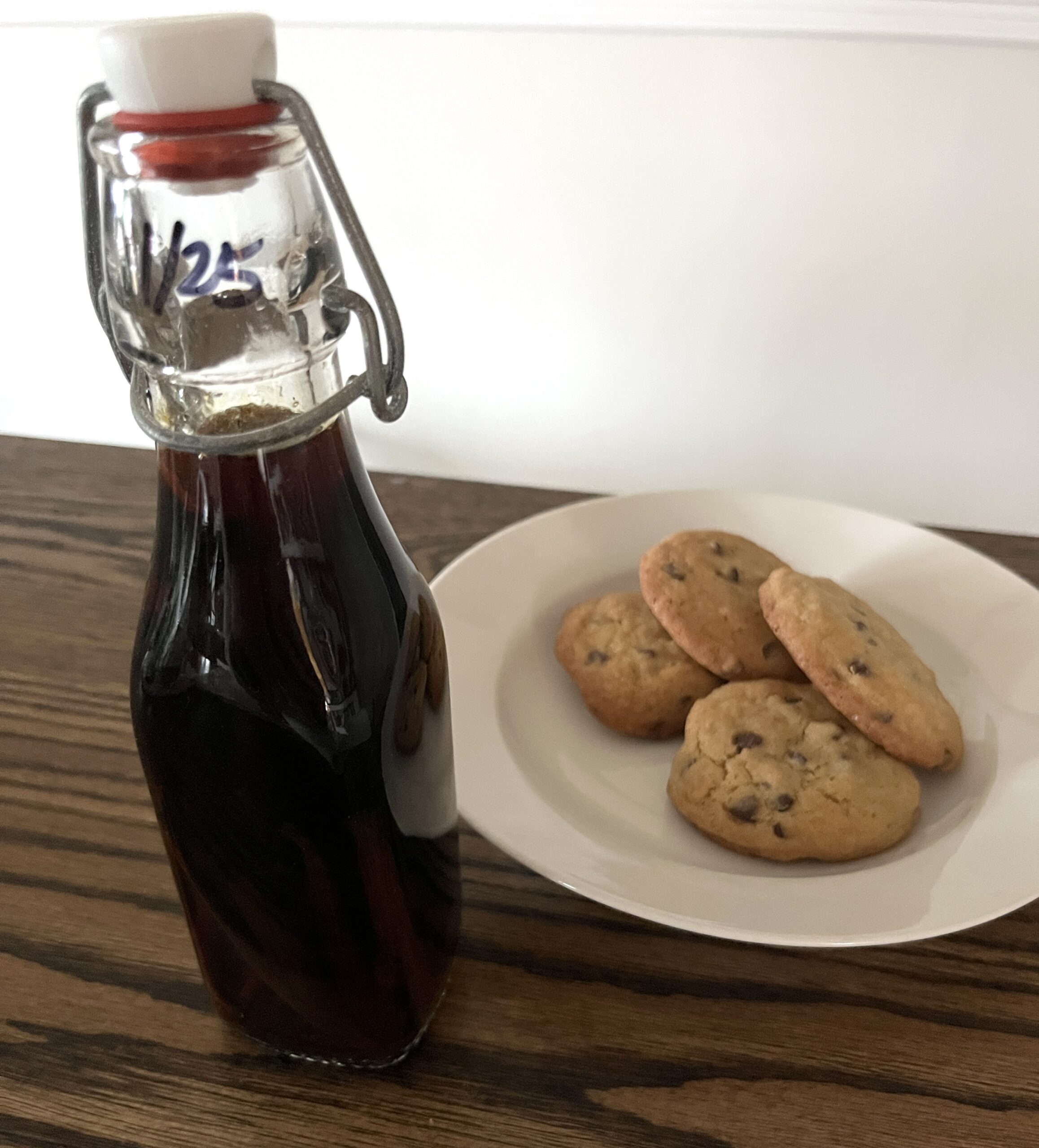 glass swing top bottle of vanilla extract sitting on wooden table in front of a small white plate of chocolate chip cookies