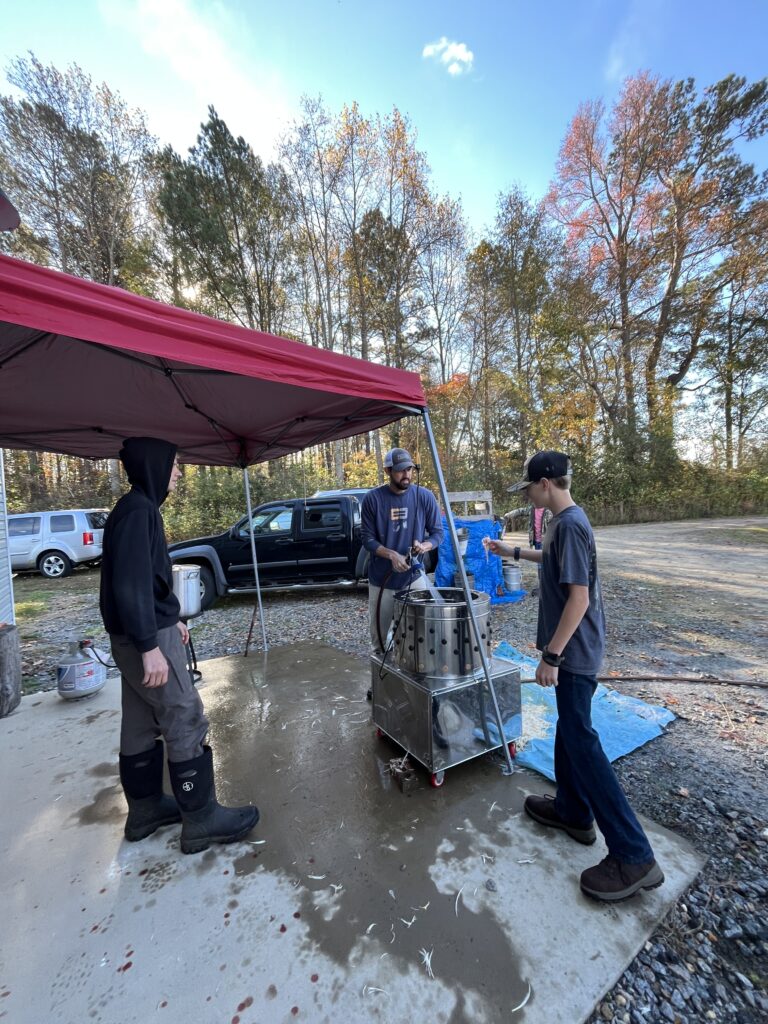 three men standing under a red tent utilizing a stainless steel chicken feather plucker machine