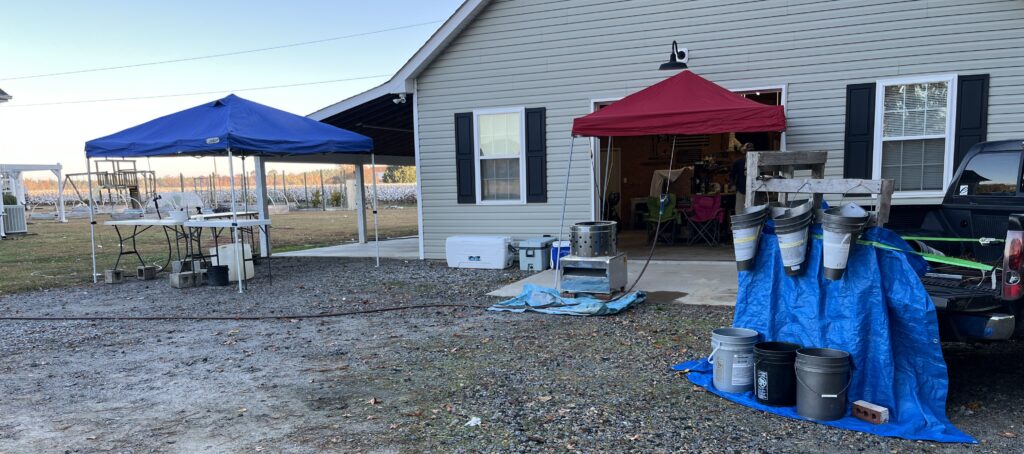 a blue and red tent set up in front of a work shop alongside a black truck bed hosting a wood pallet holding gray buckets made into cones for chicken processing stations