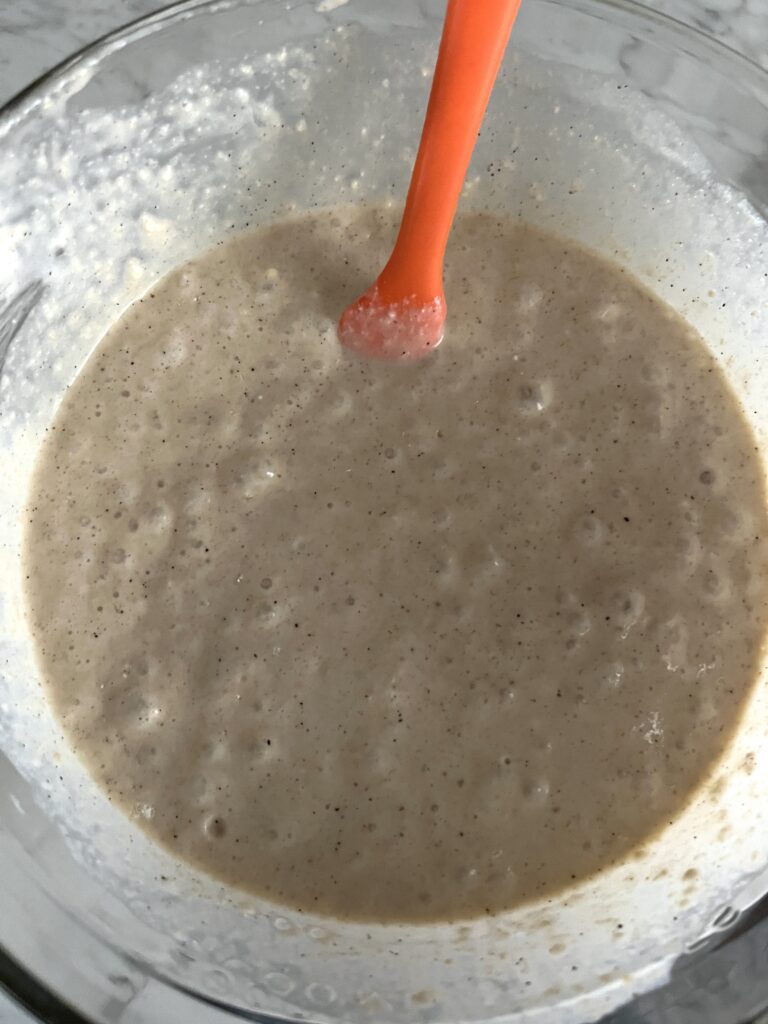 birds eye view of a bowl of freshly milled sourdough pancake batter sitting on a white counter with a orange mixing spoon in it