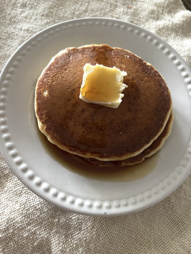 a white plate of two brown pancakes topped with a thick pat of yellow butter drizzled with maple syrup sitting on a linen napkin