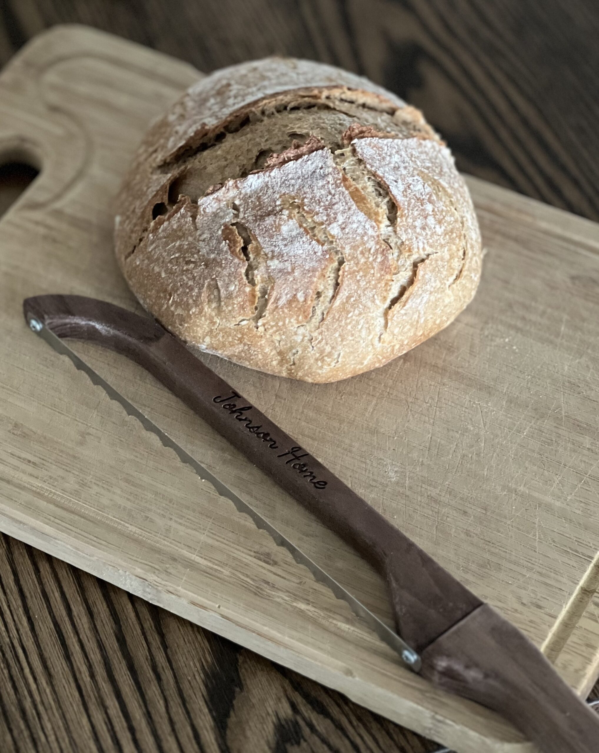 crusty round sourdough boule sitting on a wooden cutting board with a bread bow diagonally in front of the bread