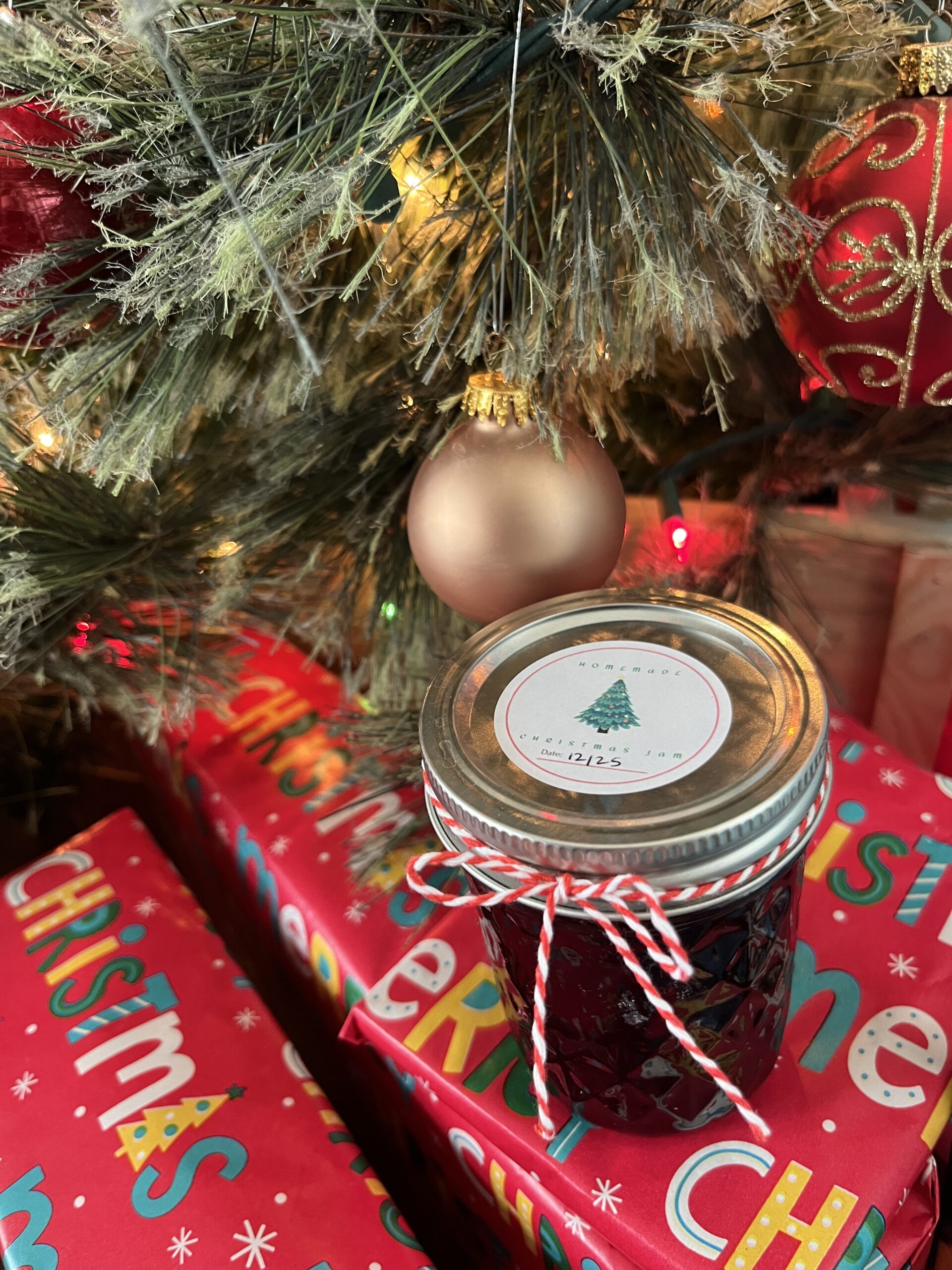 jar of Christmas jam with a red and white string tied around it sitting in front of a decorated Christmas tree on top of red wrapping paper wrapped gifts