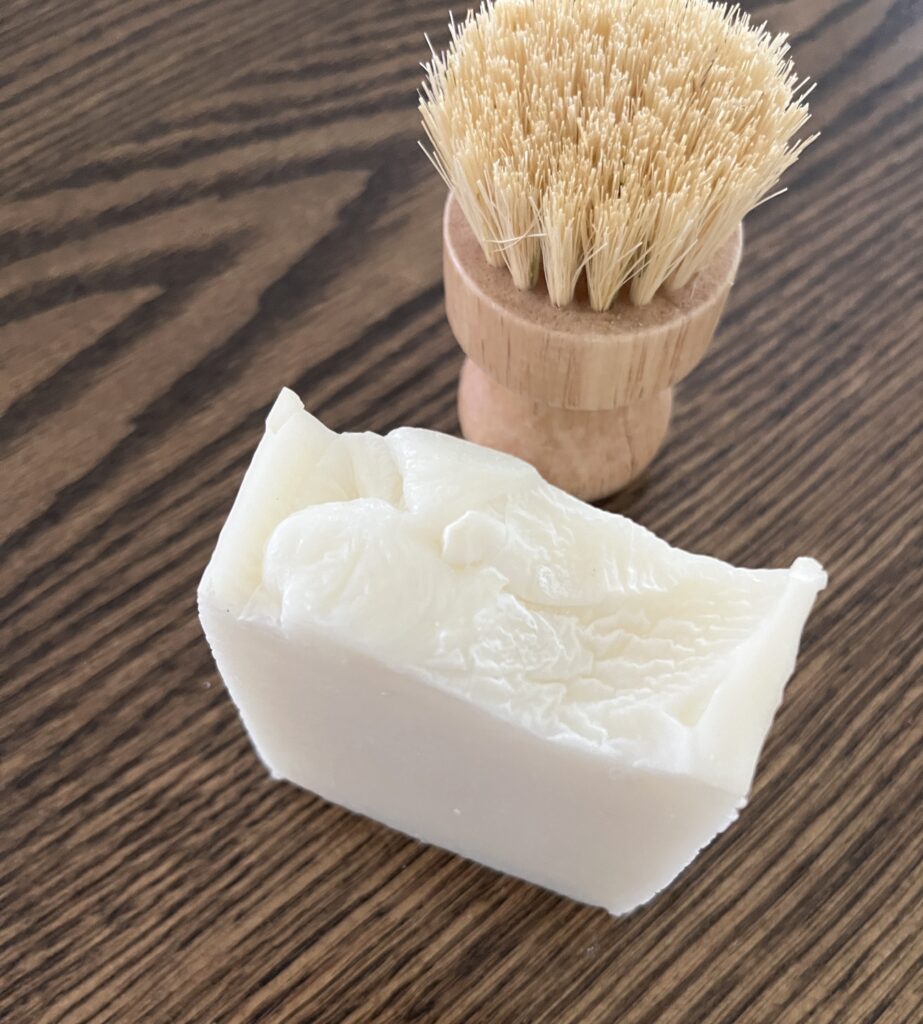 a single white bar of farmhouse dish soap sitting on a black walnut table with a bamboo dish brush behind the soap