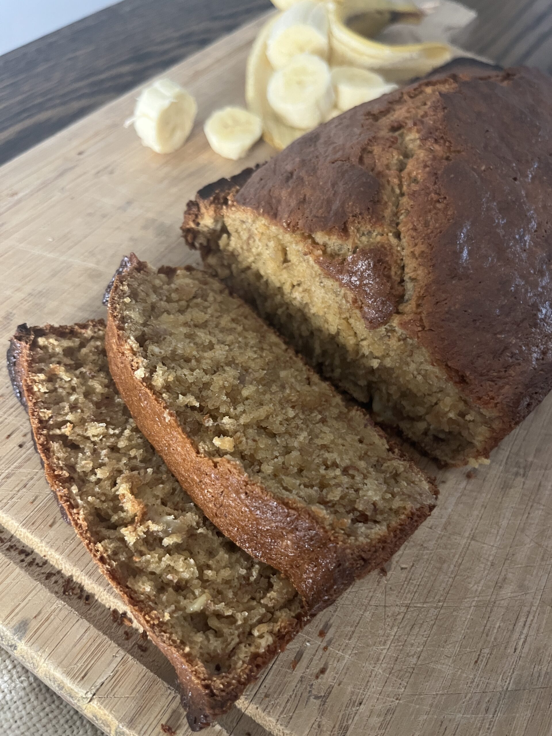 a sliced loaf of freshly milled banana bread on a wooden cutting board with sliced bananas in the background
