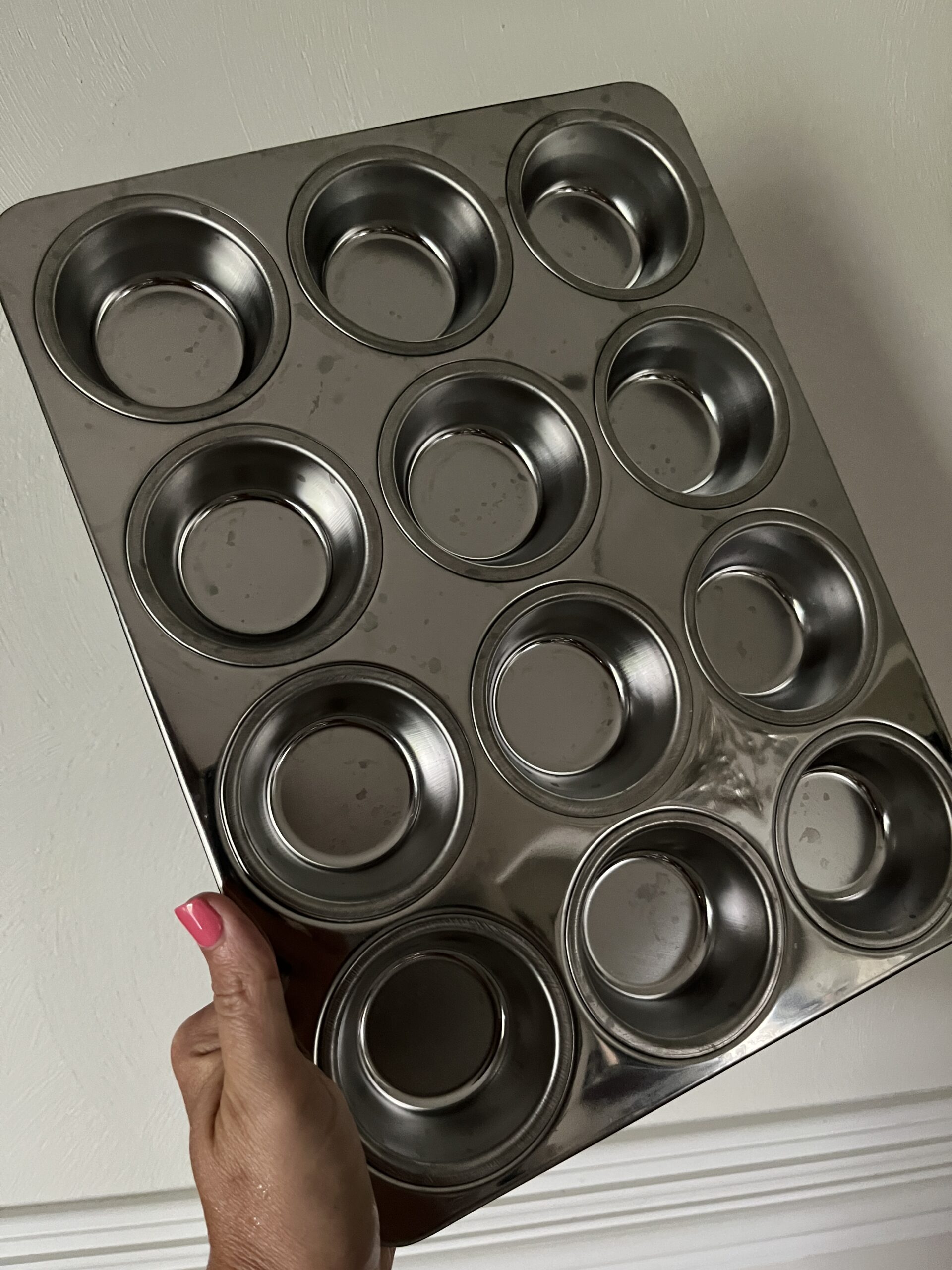 woman's hand holding up a shiny stainless steel muffin tin in front of a white wall