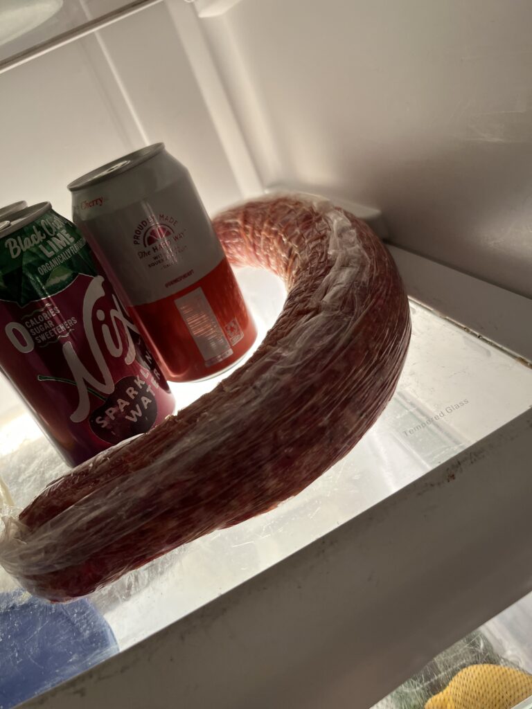 a long meat stick wrapped in plastic wrap resting inside of a refrigerator in a C curved shape around two soda cans sitting on a glass shelf