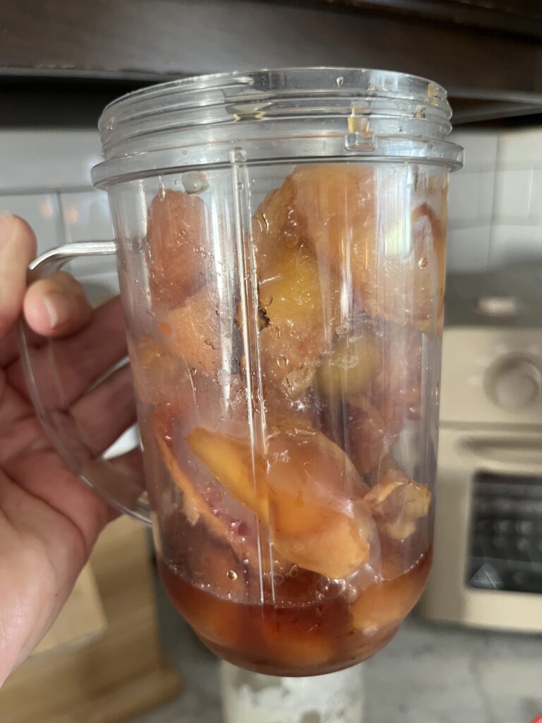 hand holding up a blender container of peach slices and maple syrup ready to be blended