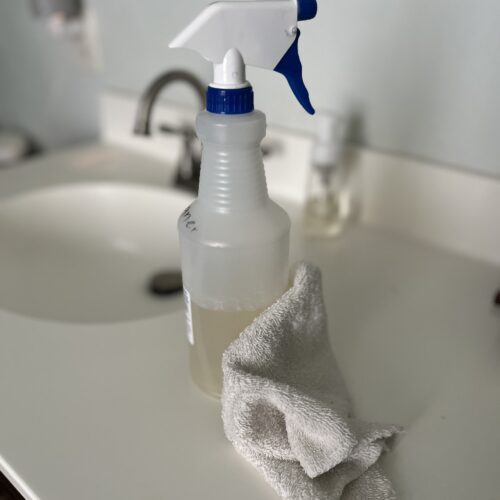 frosted plastic bottle with white and blue spray nozzle sitting on a white bathroom counter in front of a sink with a white rag on the counter in front of the all-purpose cleaner spray