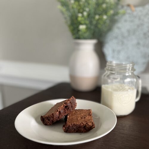 two freshly milled chocolate Brownies on a white plate sitting in front of a vase of greenery and a metal chicken decor with a mason jar of milk to the right