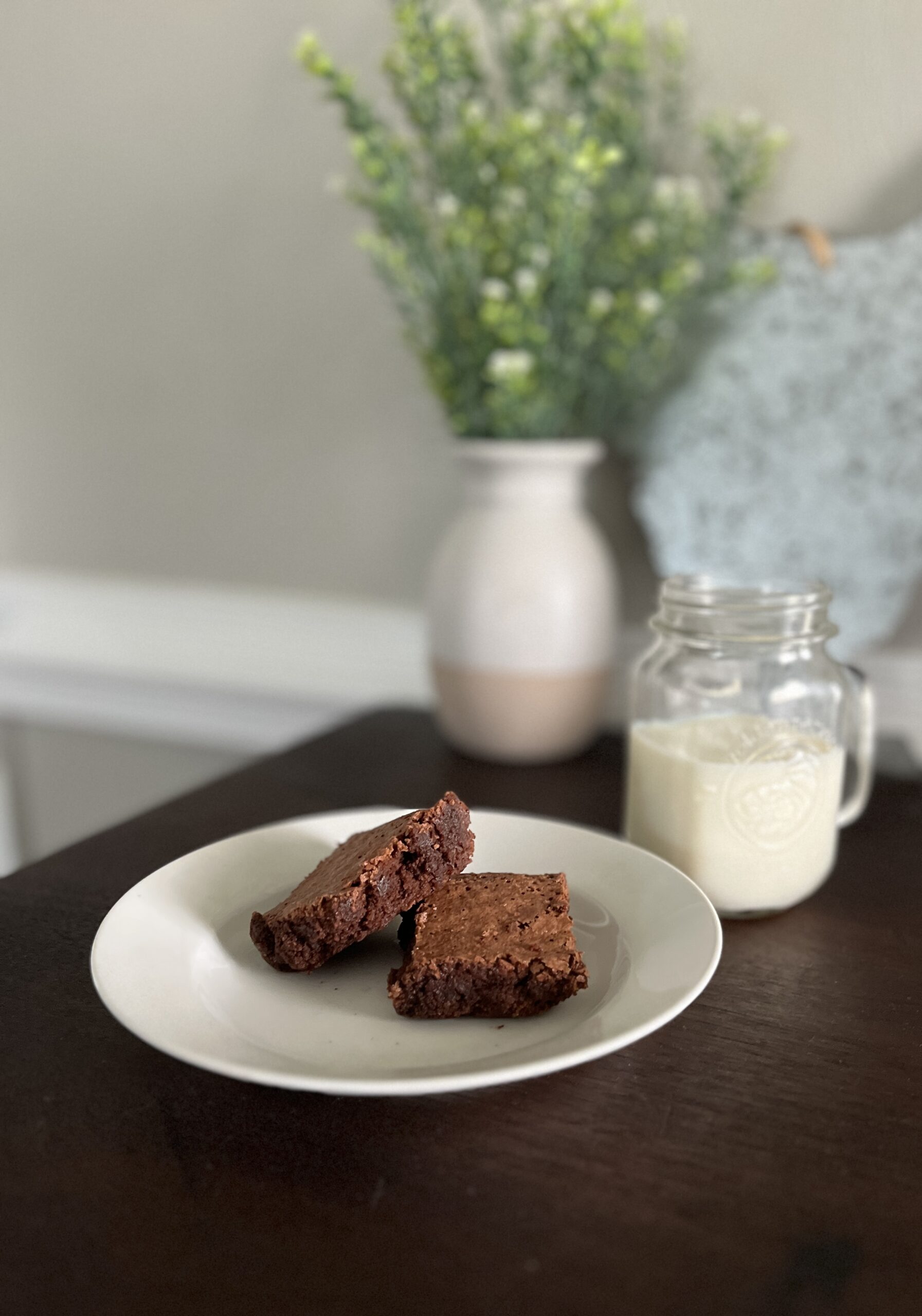 two freshly milled chocolate Brownies on a white plate sitting in front of a vase of greenery and a metal chicken decor with a mason jar of milk to the right